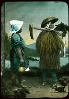 Two people in costume posing in front of painted backdrop - one in grass coat and hat, carrying farm implement over shoulder and holding vegetables; the other, a woman in a blue dress with a red (over mantel), legs bare to the knees, wearing a white kerch